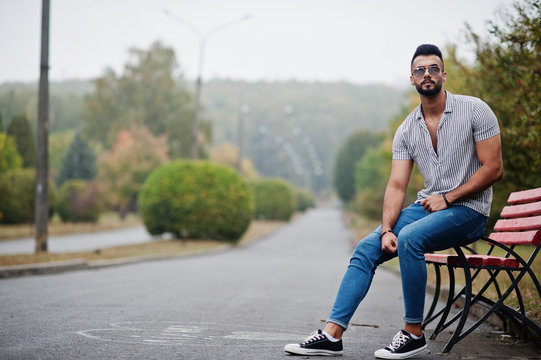 Fashionable Tall Arab Beard Man Wear On Shirt, Jeans And Sunglasses Posed On Park At Bench.