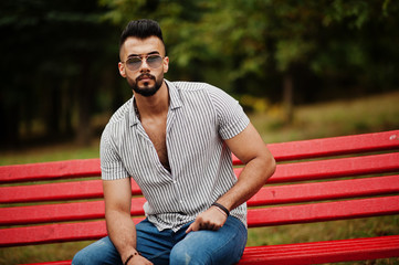 Obraz premium Fashionable tall arab beard man wear on shirt, jeans and sunglasses sitting on red bench at park.