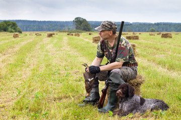 the hunter sits on the mown hay and holds a downed grouse in his hands