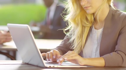Medium shot of young blonde woman sitting at table outdoors and typing something on laptop computer keyboard - Powered by Adobe