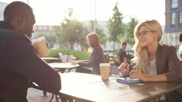 Medium shot of young blonde businesswoman in glasses sitting at table in outdoor cafe or coworking space and talking to black male colleague over cup of coffee