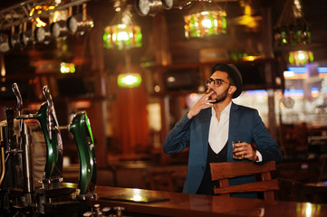 Handsome well-dressed arabian man with glass of whiskey and cigar posed at pub.