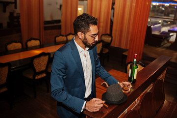 Handsome well-dressed arabian man with glass of whiskey and cigar posed at pub.