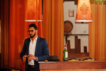 Handsome well-dressed arabian man with glass of whiskey and cigar posed at pub.