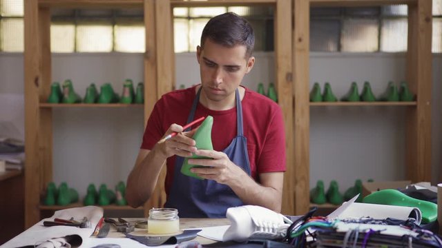 Medium shot of skilled young shoemaker applying glue to plastic last with brush while making standing at table in workshop