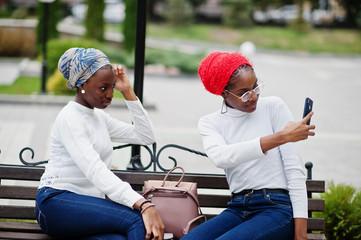 Naklejka premium Two young modern fashionable, attractive, tall and slim african muslim womans in hijab or turban head scarf posed together with mobile phones at hands.