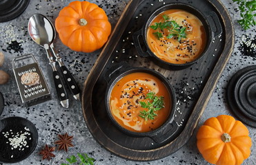 Pumpkin soup with cream in a cast iron pot. Homemade pumpkin soup served with cream, butter, sesame and parsley on a grey concrete background