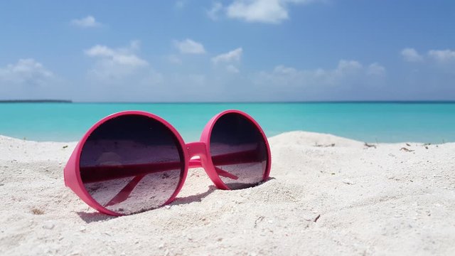 A pair of pink sunglasses with purple lenses sits on top of bright white sand on a tropical beach in Thailand