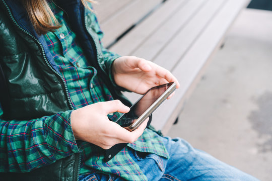 A Boy With Long Hair In A Green Plaid Shirt And Vest Is Sitting On The Bus Stop Bench With A Broken Phone In His Hands. Concept Of Gadget Addiction And Modern Technology. Copy Space