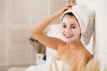 smiling woman with towel is applying mask on her face and looking at the camera at home. Skin care. space for text