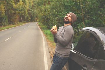Young guy eating a burger near a car on an empty road. Food on the trip. Food on the go. Autumn travel. Fast food.
