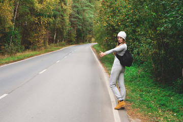 A young and beautiful girl votes on the road. Autumn road. Catch a car on an empty road. Hitchhiking. Free travel by car.