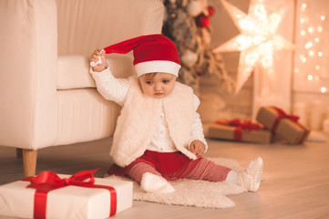 Obraz na płótnie Canvas Cute baby girl 1-2 year old wearing santa claus har and suit sitting on floor in room over Christmas tree with glowing lights and presents close up. Winter holiday season. Childhood.