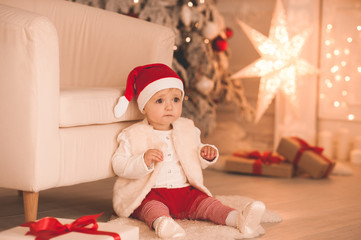 Obraz na płótnie Canvas Sweet baby girl 1-2 year old sitting on floor wearing santa claus hat and suit in room over Christmas tree and glowing lights close up. Winter holiday season. Happy New Year. Childhood.