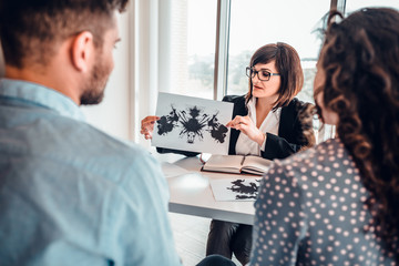 Fototapeta premium Family at psychotherapy session. Mature psychotherapist showing sheets with Rorschach test to young married couple, making psycho analysis. View from behind patients. Relationship psychology concept