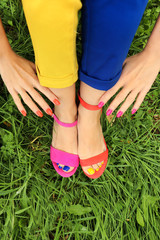 Colorful bright pedicure in different pink and orange sandals and different blue and yellow pants on a background of green grass.Fashionable summer nail art.