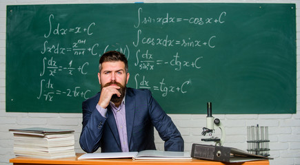 Thinking on serious problem. Serious teacher touching beard by hand. Bearded man with serious face sit at teachers table. Hipster having serious look in school. Solving problem. Dilemma