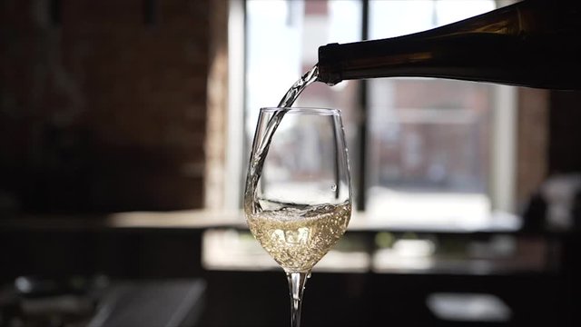  White wine is poured into a glass from a bottle against the background of a window in a restaurant on a silhouette