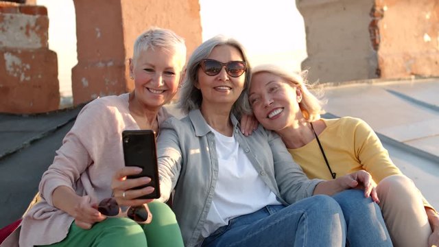 Medium shot of three middle-aged women sitting together on old roof, hugging and making selfie