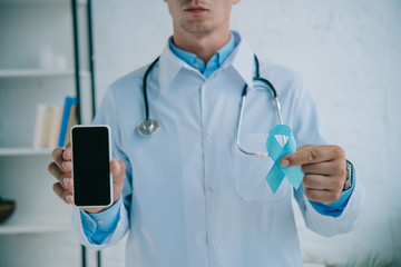 cropped view of doctor holding blue awareness ribbon and smartphone with blank screen