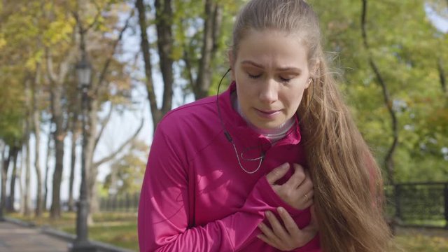 Young Caucasian woman in pink sportswear stopping during the run because of chest pain. Female runner feeling bad during the morning training in the autumn park. Sports and healthcare concept.