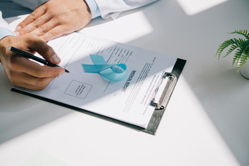 cropped view of doctor writing in medical report near blue awareness ribbon