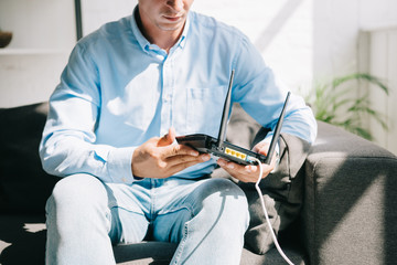 cropped view of businessman sitting on sofa and holding plugged router - obrazy, fototapety, plakaty