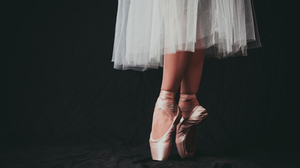 Close-up of dancing legs of ballerina wearing white pointe on a black background. - obrazy, fototapety, plakaty