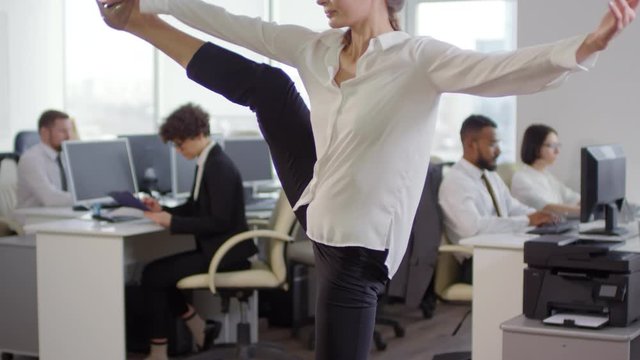 Tilt up of young Caucasian businesswoman stretching her legs and arms in office while her coworkers working on PCs