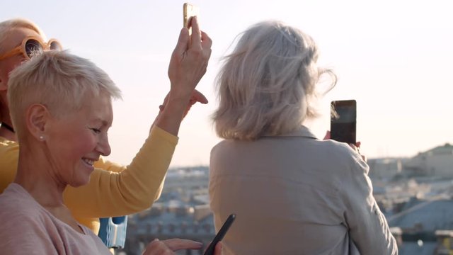 Close up of three happy middle-aged women wearing casual clothes standing on roof of building with smartphones in their hands and making photos