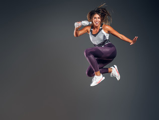 Fototapeta na wymiar Exited cheerful woman is jumping at studio while holding the bottle of water over grey background.