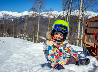 Fototapeta na wymiar Portrait of little boy snowboarder sitting on the snow during the winter vacation at ski resort, on background beautiful landscape of snowy high mountains in sunny day