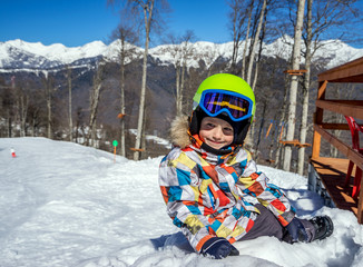 Portrait of little boy snowboarder sitting on the snow during the winter vacation at ski resort, on...