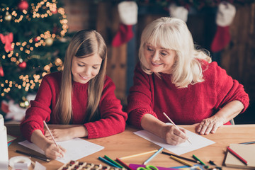 Fototapeta premium Portrait of nice attractive lovely inspired cheerful granny pre-teen grandchild writing festal wish letter to Santa Christmastime at decorated industrial brick wood loft style lights interior house