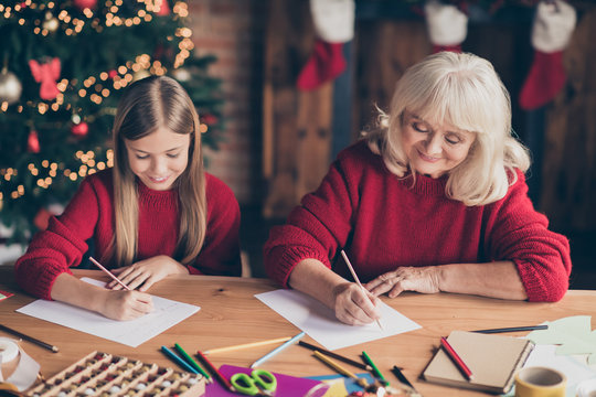 Portrait of nice attractive cheerful cheery granny pre-teen grandchild writing wish want list December on table at decorated industrial brick wood loft style lights interior house