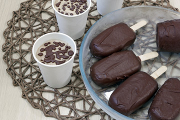 Delicious homemade diet ice cream. Raw vegan dessert with natural chocolate. Small portions in white paper cups and popsicle on a stick. Glass plate on wicker tablecloth on the kitchen table