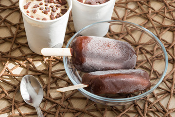 Healthy vegan homemade dessert on wicker tablecloth. Raw banana ice cream coated with natural chocolate in a white paper cup and chocolate popsicles on sticks in glass bowl, with spoon. Right eating