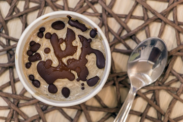 Raw banana ice cream with coconut cream, coated with natural chocolate, in a white paper cup with spoon on brown wicker tablecloth. Delicious healthy vegan sweet dessert. Top view. Close-up