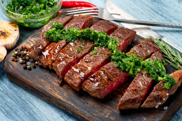 Sliced juicy beef steak with chimichurri sauce and spices on wooden board.