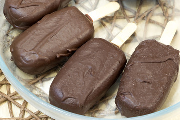 Fresh raw popsicles on sticks coated with chocolate glaze lay in glass transparent bowl on wicker tablecloth. Delicious vegan homemade dessert. Top angle view. Close-up. Selective focus. Healthy meal