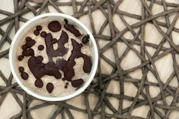 Top view healthy vegan sweet frozen dessert. Raw banana ice cream with coconut cream coated with natural chocolate droplets in a white paper cup on table covered with wicker tablecloth. Copy space