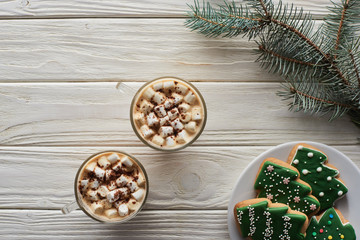 top view of cacao with marshmallow and cacao powder in mugs near Christmas cookies and fir branch on white wooden table