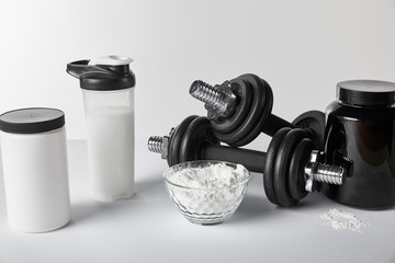 Obraz na płótnie Canvas jars and sports bottle near bowl with protein powder and dumbbells on white