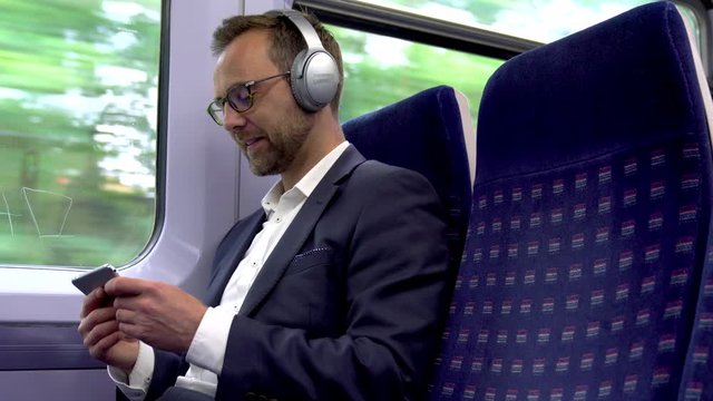 Mature Businessman On Train Wearing Wireless Headphones Streaming Online Content To Mobile Phone