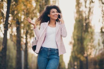 Photo of amazing pretty dark skin lady spending time walk park after university lectures hold telephone speak best friend autumn weather wear jacket jeans outdoors