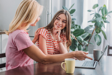 mother pointing at laptop while touching shoulder of young, smiling daughter