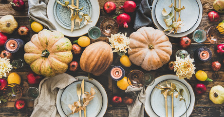 Fall table setting for Thanksgiving day or family dinner. Flat-lay plates, silverware, floral and fruit decoration, candle and pumpkins over rustic wooden table background, top view, wide composition