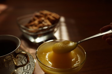 Honey in a glass bowl with a spoon in hand, a cup of tea on a saucer, cookies in a vase on the brown sunny table.