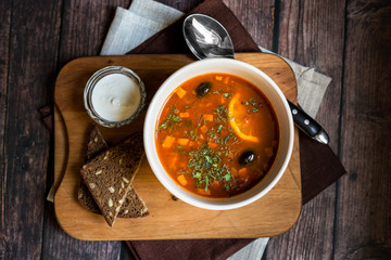Solyanka soup in a white bowl on the dark rustic wooden table with slices of dark bread and sour cream