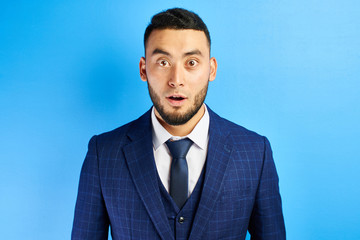 Portrait of excited Kazakh Asian man with surprised open mouth and bulging eyes in a blue suit with a tie isolated in studio